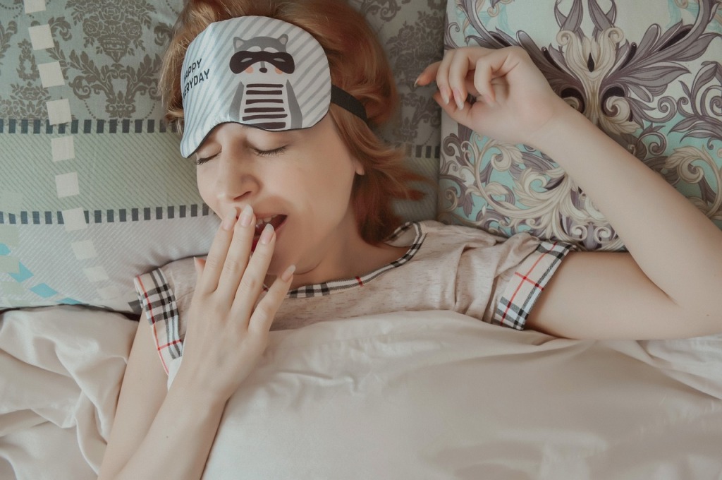 A woman is lying in bed yawning. She is wearing a raccoon sleep mask over her forehead.